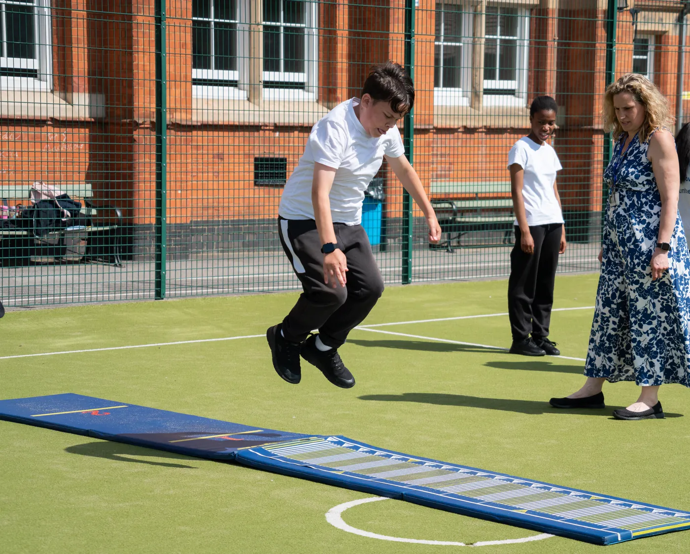 A student jumping from a long-jump board on the playground, a classmate and a member of staff watching from beside the run-up.
