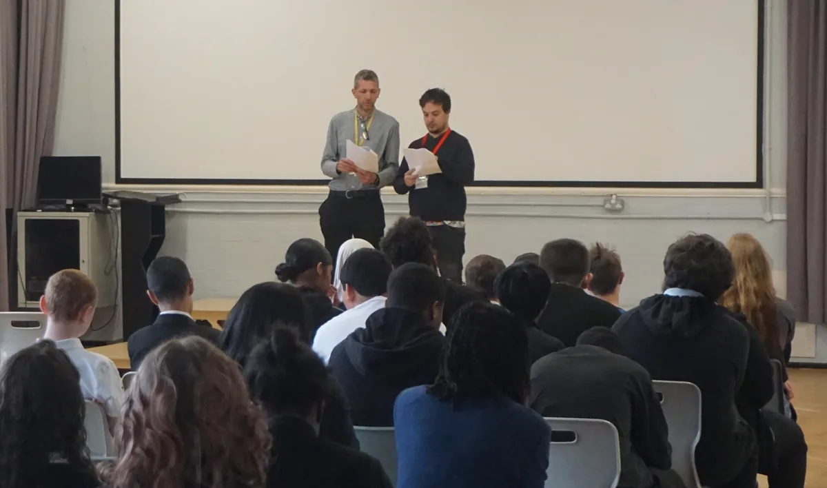 Anton standing with a member of staff at the front of the hall, addressing seated students.