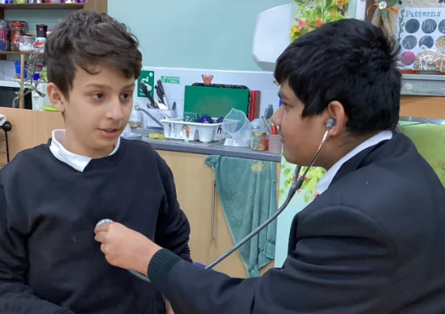 A student listening to another student’s chest with a stethoscope in a classroom.
