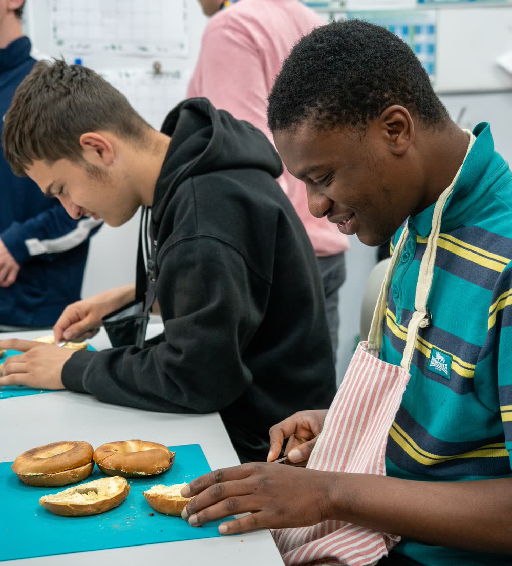 Two students at a food preparation bench, one in a striped apron spreading butter on a bagel.