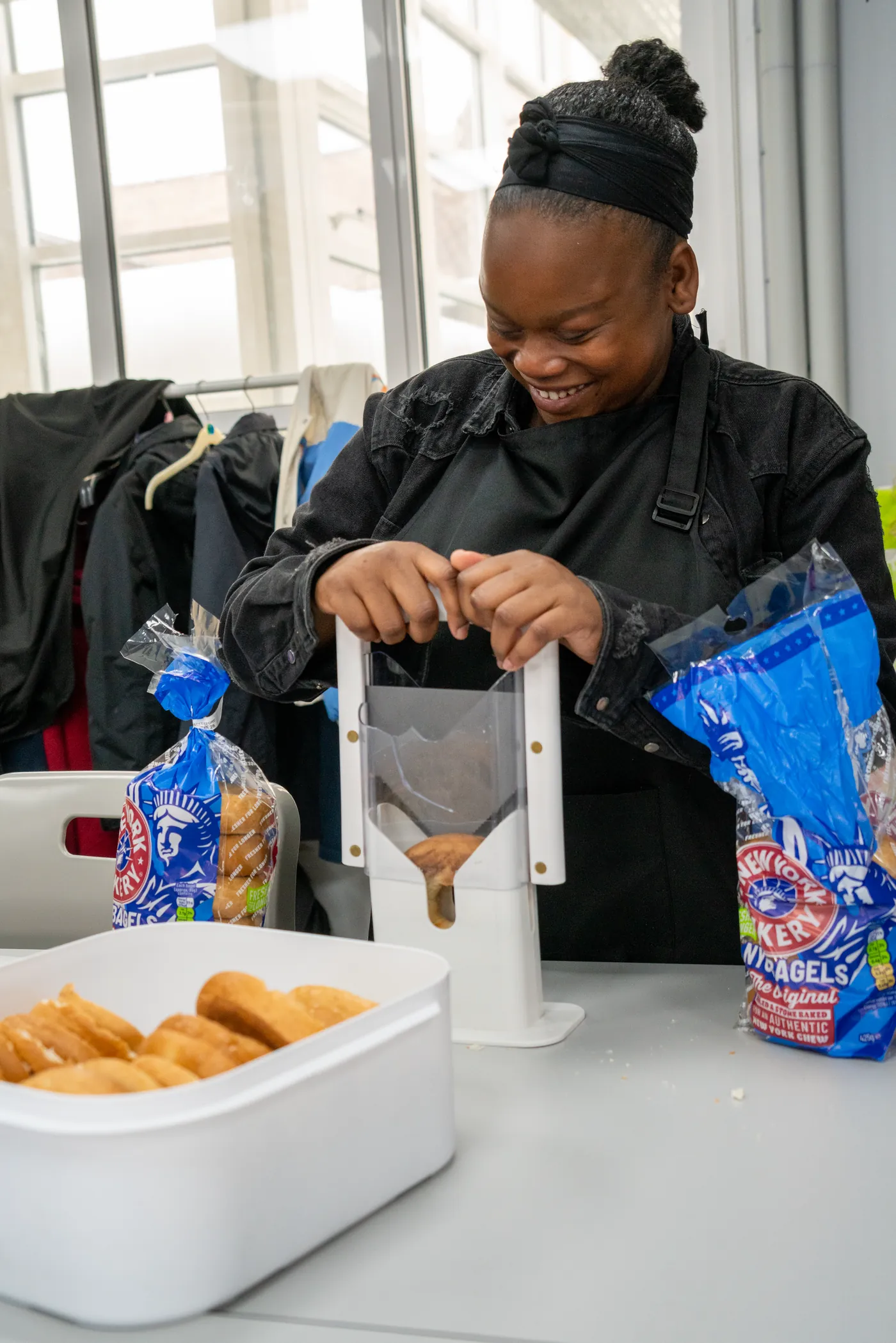 A student in an apron smiling as they slice bagels on a food preparation table, a tub of sliced bagels beside them.