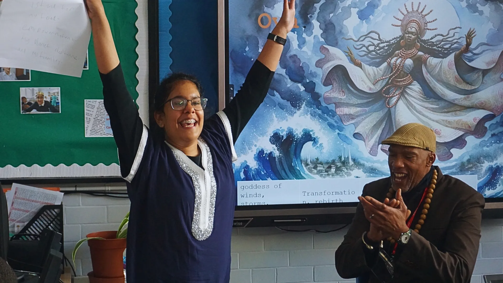 A student standing with both arms raised beside a visiting speaker who is applauding, in front of a classroom display of artwork.