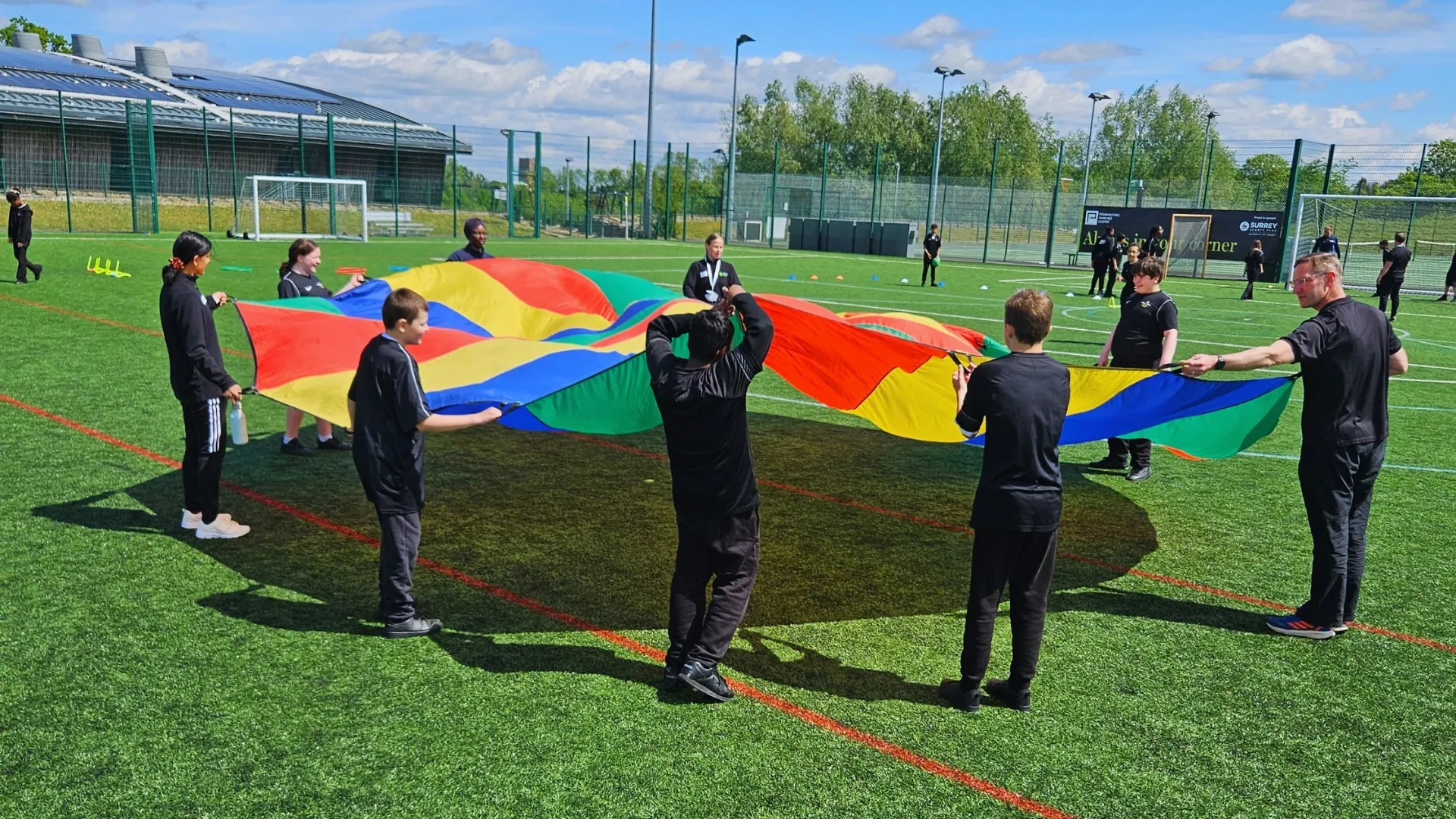Students and staff on an all-weather pitch on a sunny day, holding the edges of a rainbow parachute between them.