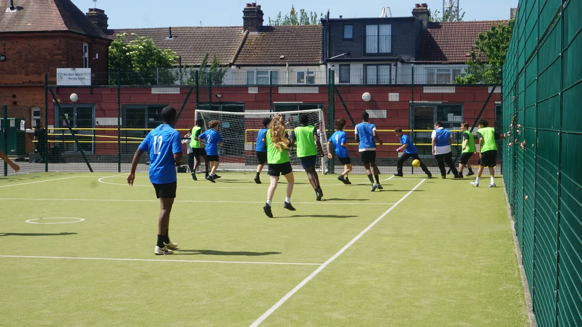 A football match on the all-weather pitch, students in blue and green bibs.