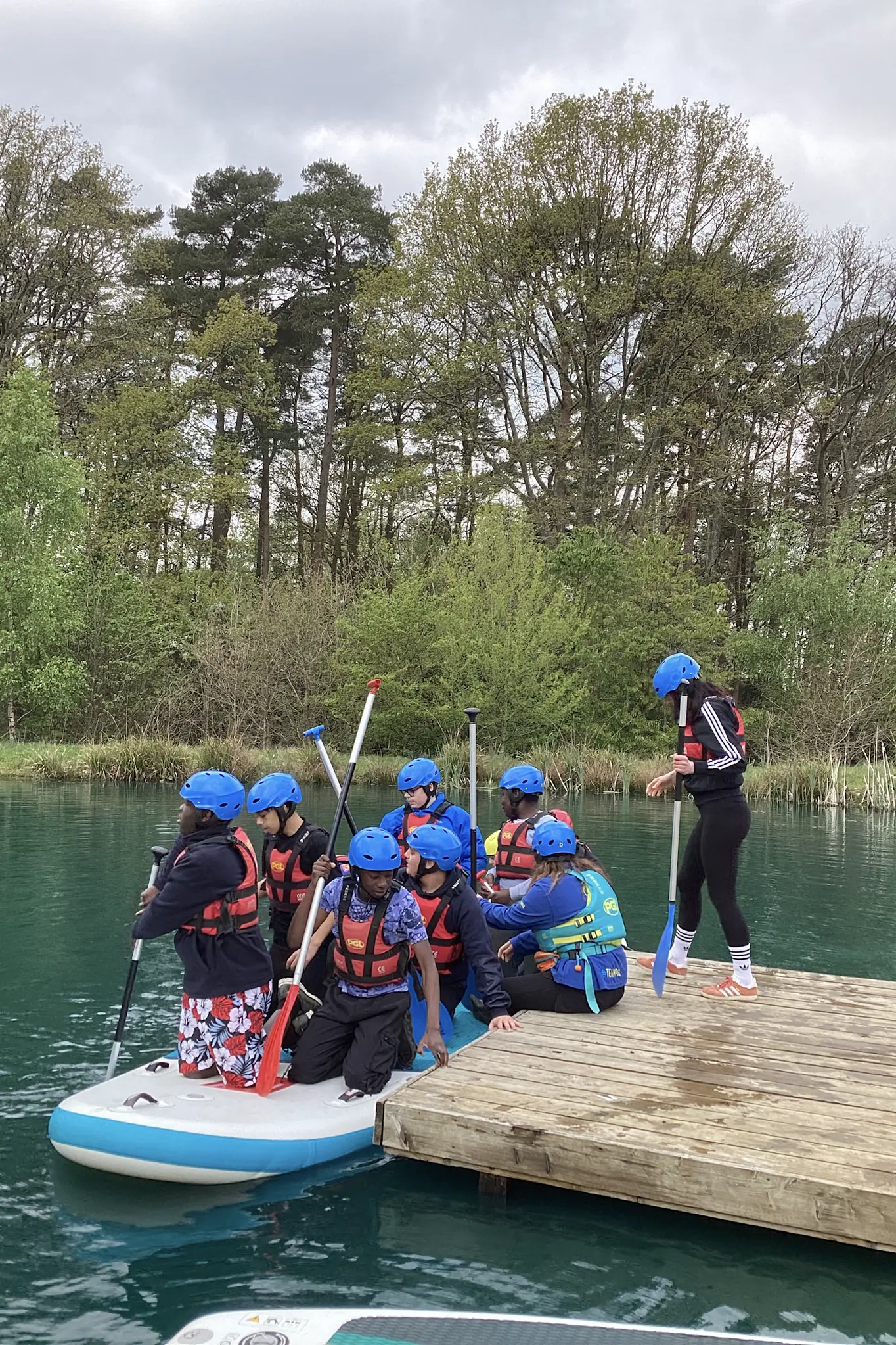 Students in helmets and buoyancy aids on a paddleboard beside a wooden jetty on a lake, woodland behind them.