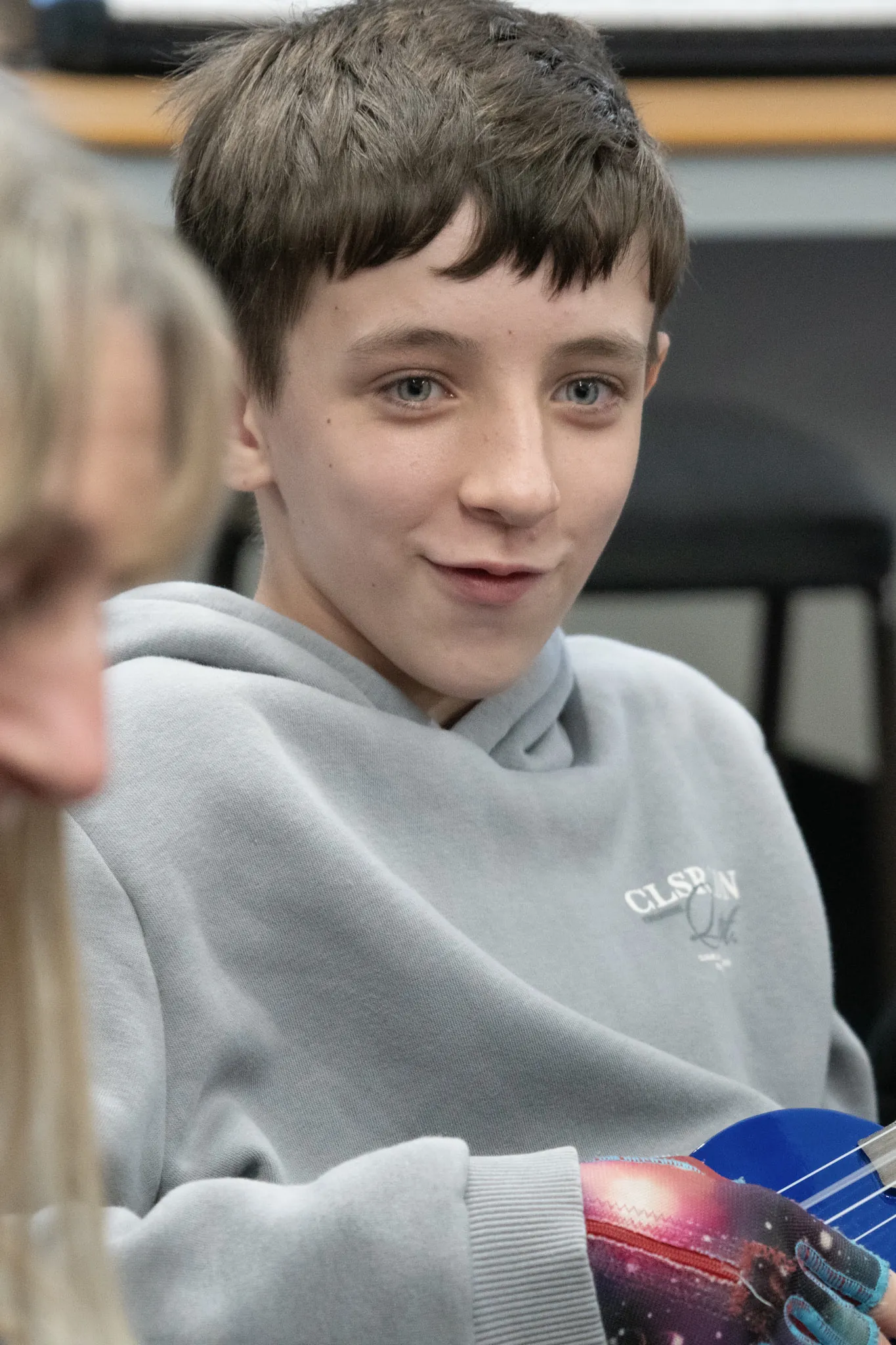 A student in a grey hoodie holding a ukulele, turning to smile at someone beside them.