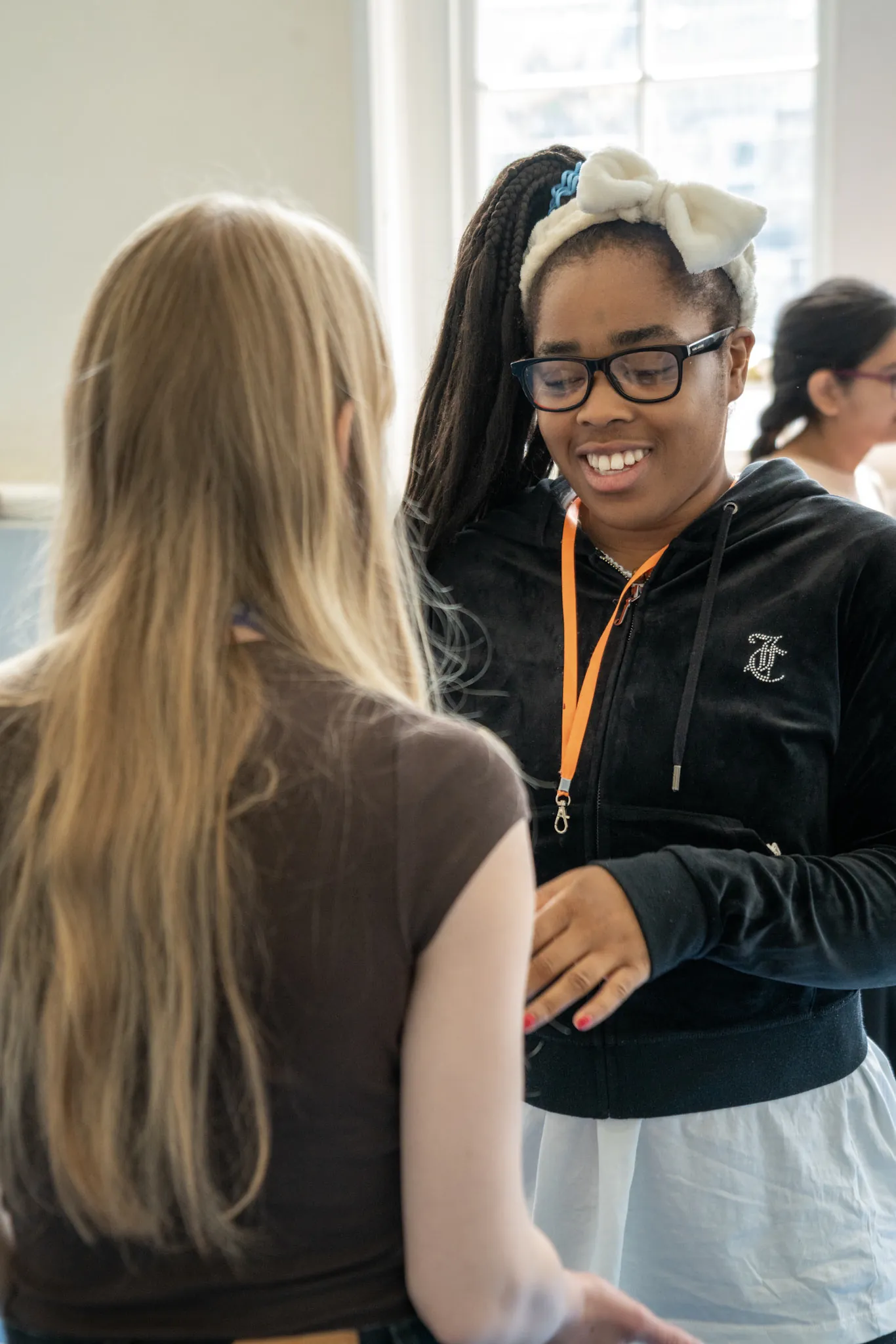 Two students standing face to face in a sunlit classroom, one of them smiling as they talk.