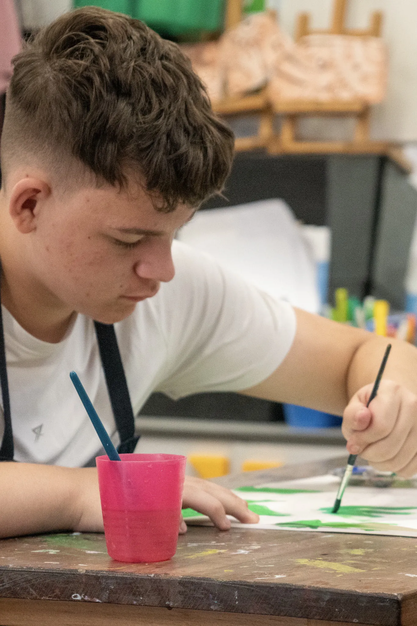 A student in an apron leaning over a workbench, painting in green on a sheet of paper, a pink water pot beside them.