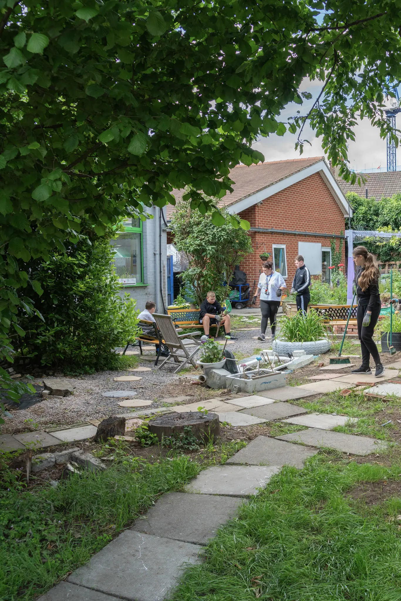 The school garden on a summer day: stepping stones across the lawn, raised beds, and students and staff by a brick outbuilding under a tree canopy.