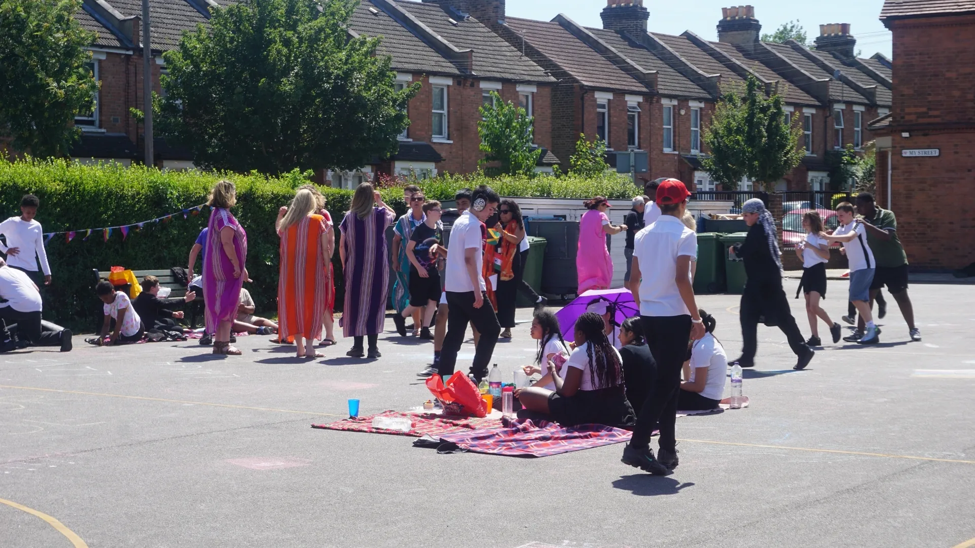 Students in cultural dress standing and sitting on the playground on a sunny day, some on a picnic blanket, with bunting strung along the fence behind.