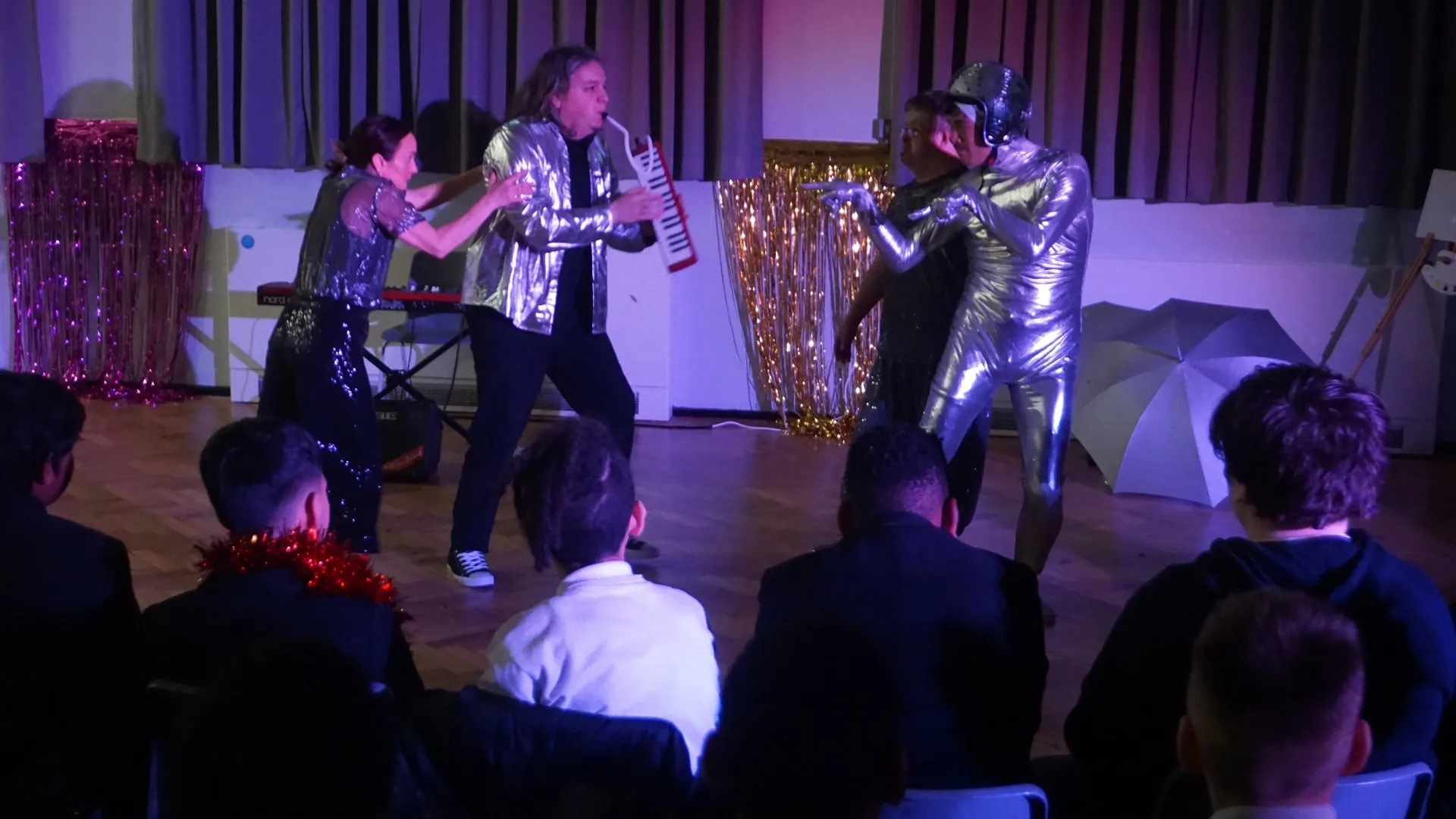 Students performing on stage in silver costumes under coloured lighting, an audience seated in front.