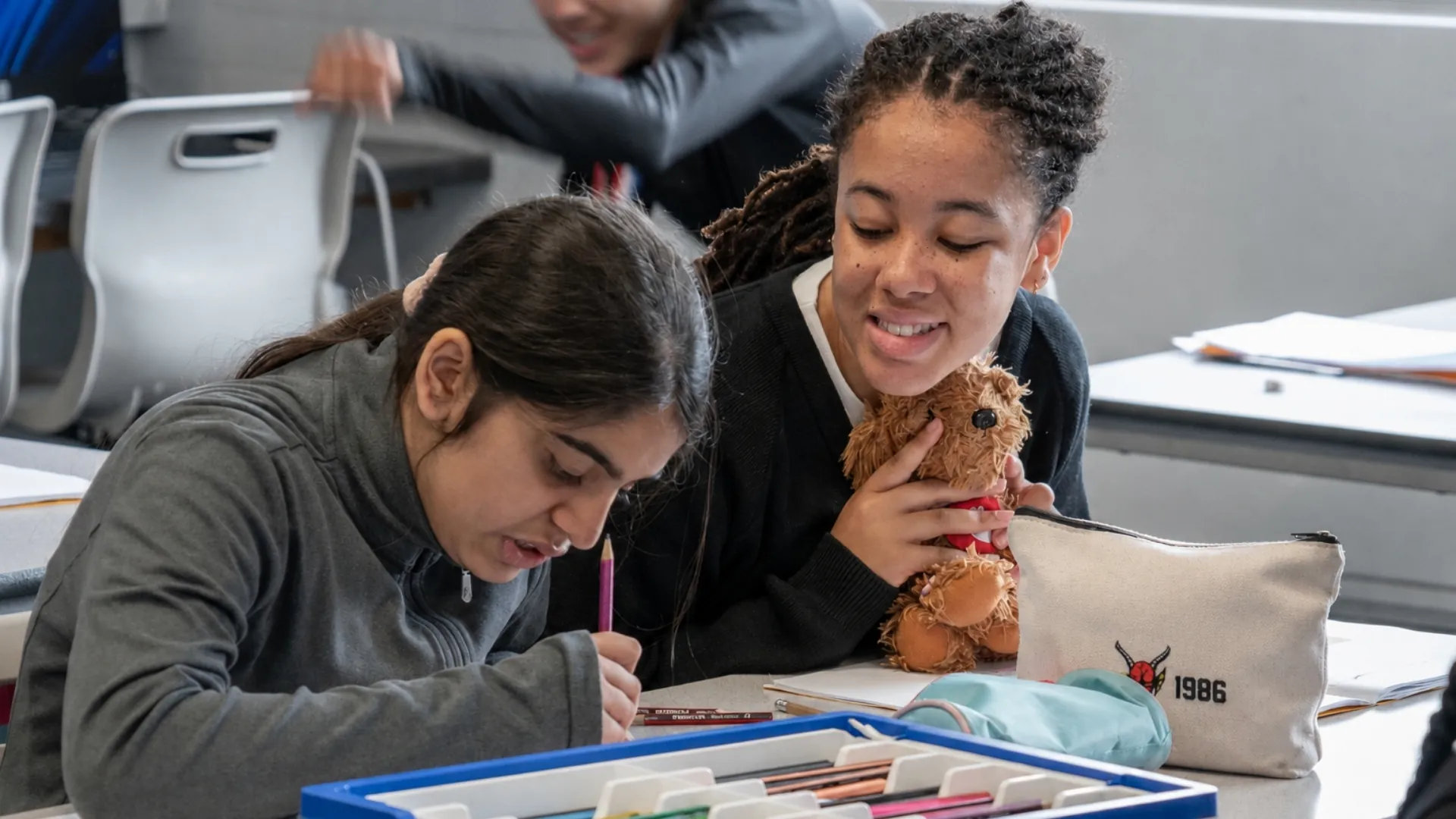 Two students working side by side at a classroom table, one writing with a coloured pencil while the other holds a soft toy.