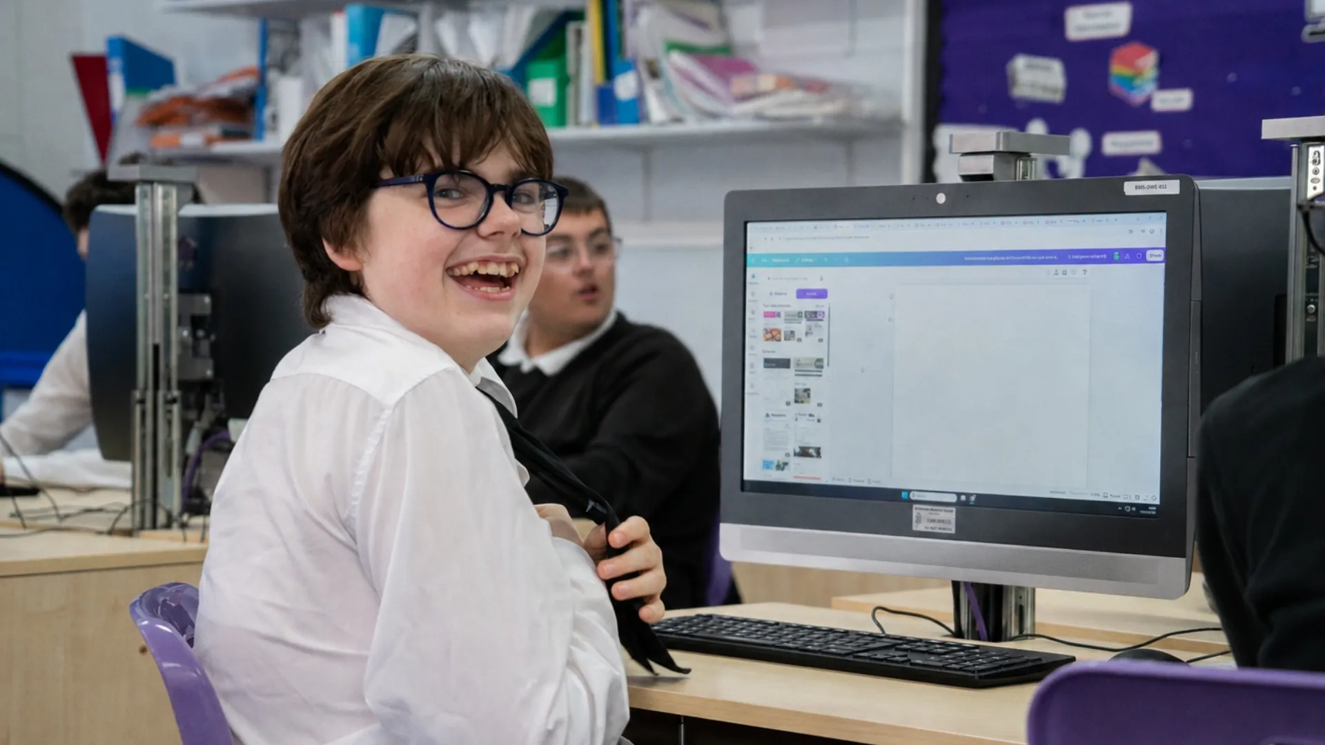 A student turning from a desktop computer to smile at the camera, editing software open on the screen behind.