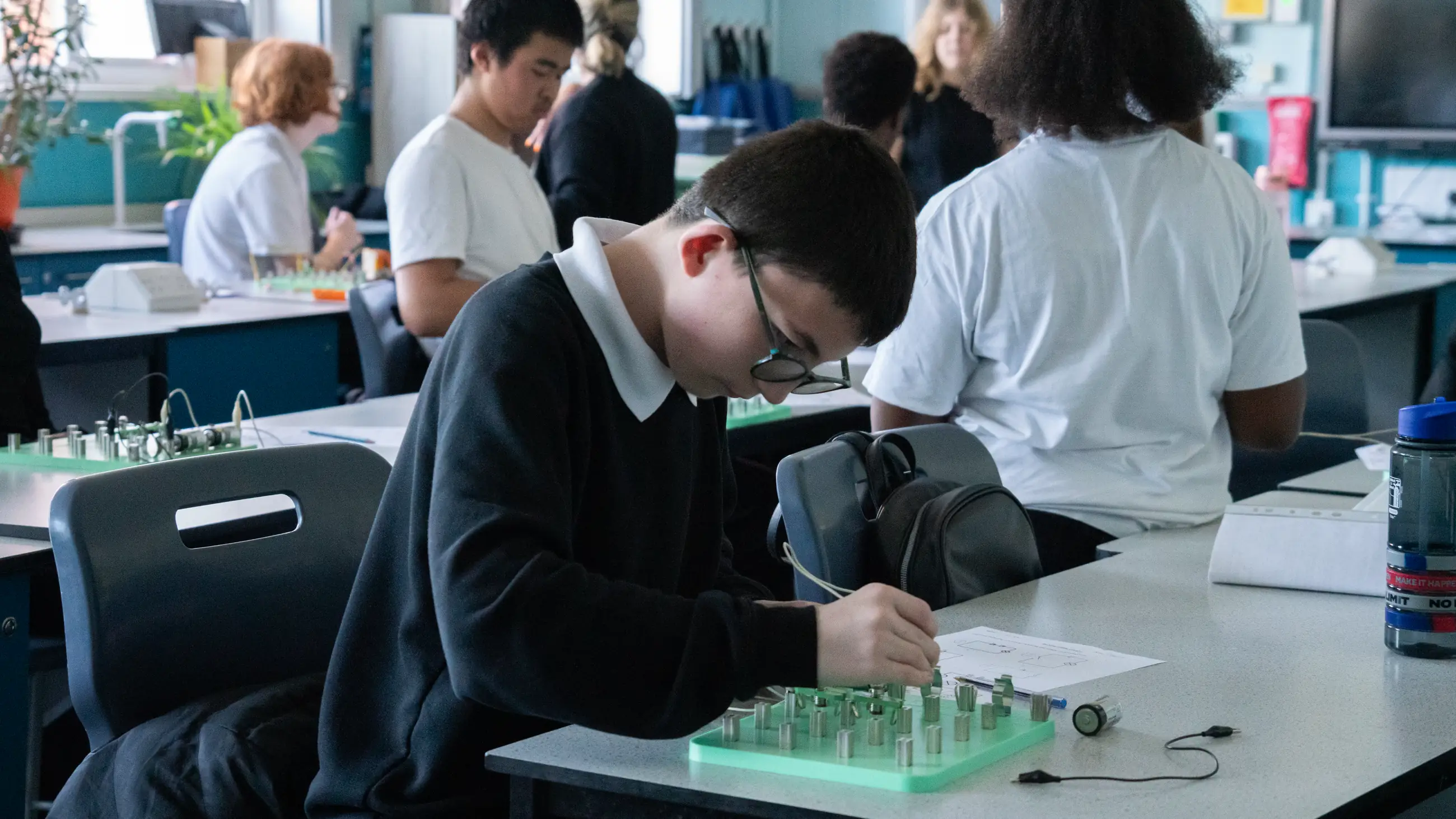 A student writing at a laboratory bench, a green component board and equipment in front of them, classmates working behind.