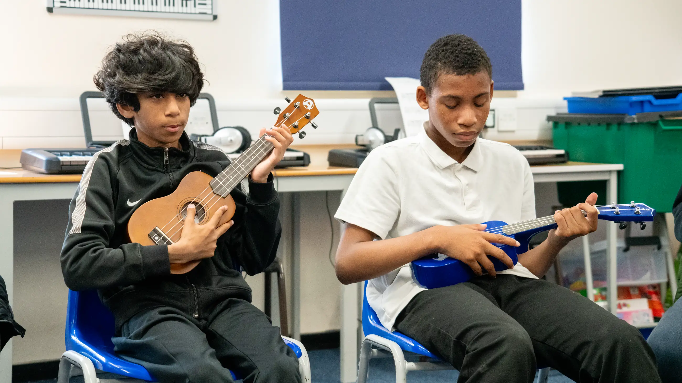 Two students sitting side by side playing ukuleles in a classroom.