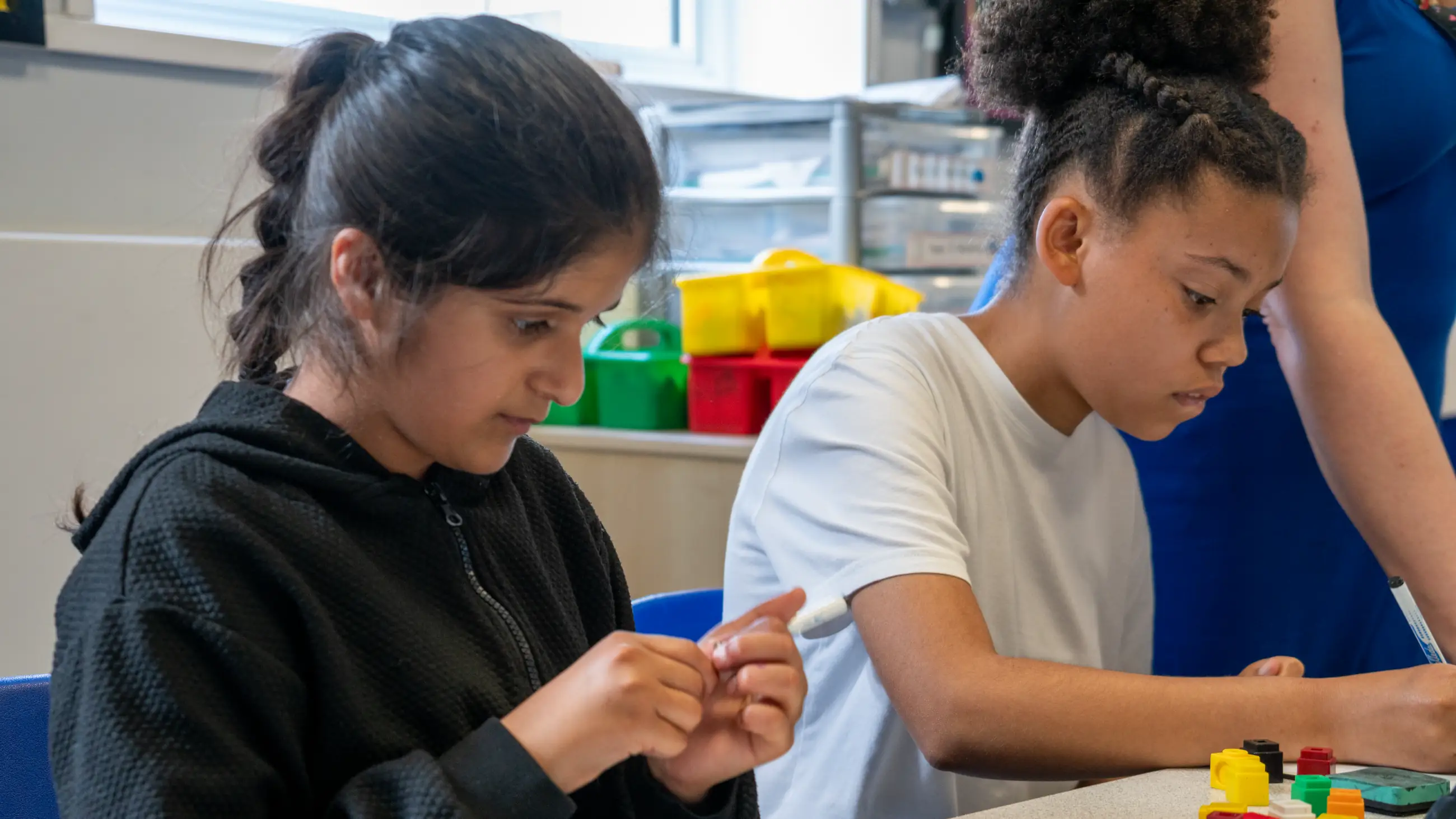 Two students working side by side at a classroom table, coloured counters and trays beside them.