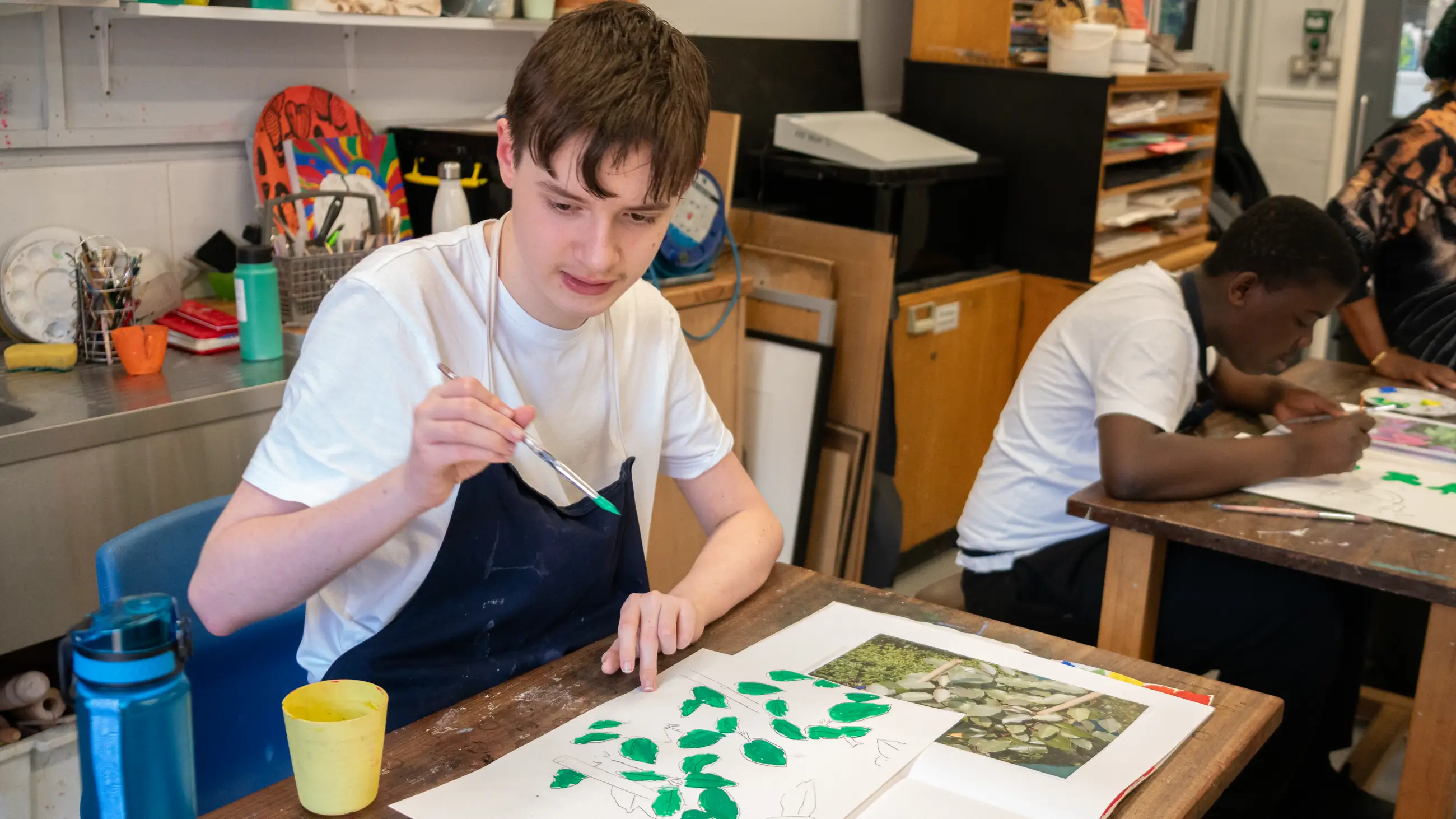 A student painting green leaf shapes onto paper at a workroom table, classmates working behind.