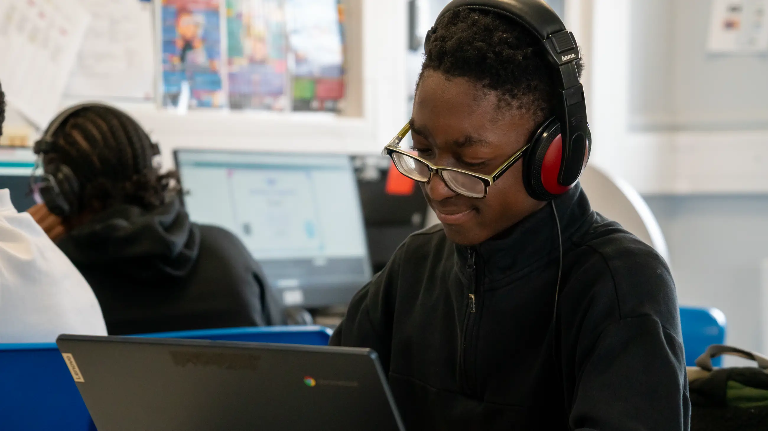 A student in headphones smiling down at a laptop in a computer room, a classmate working behind.