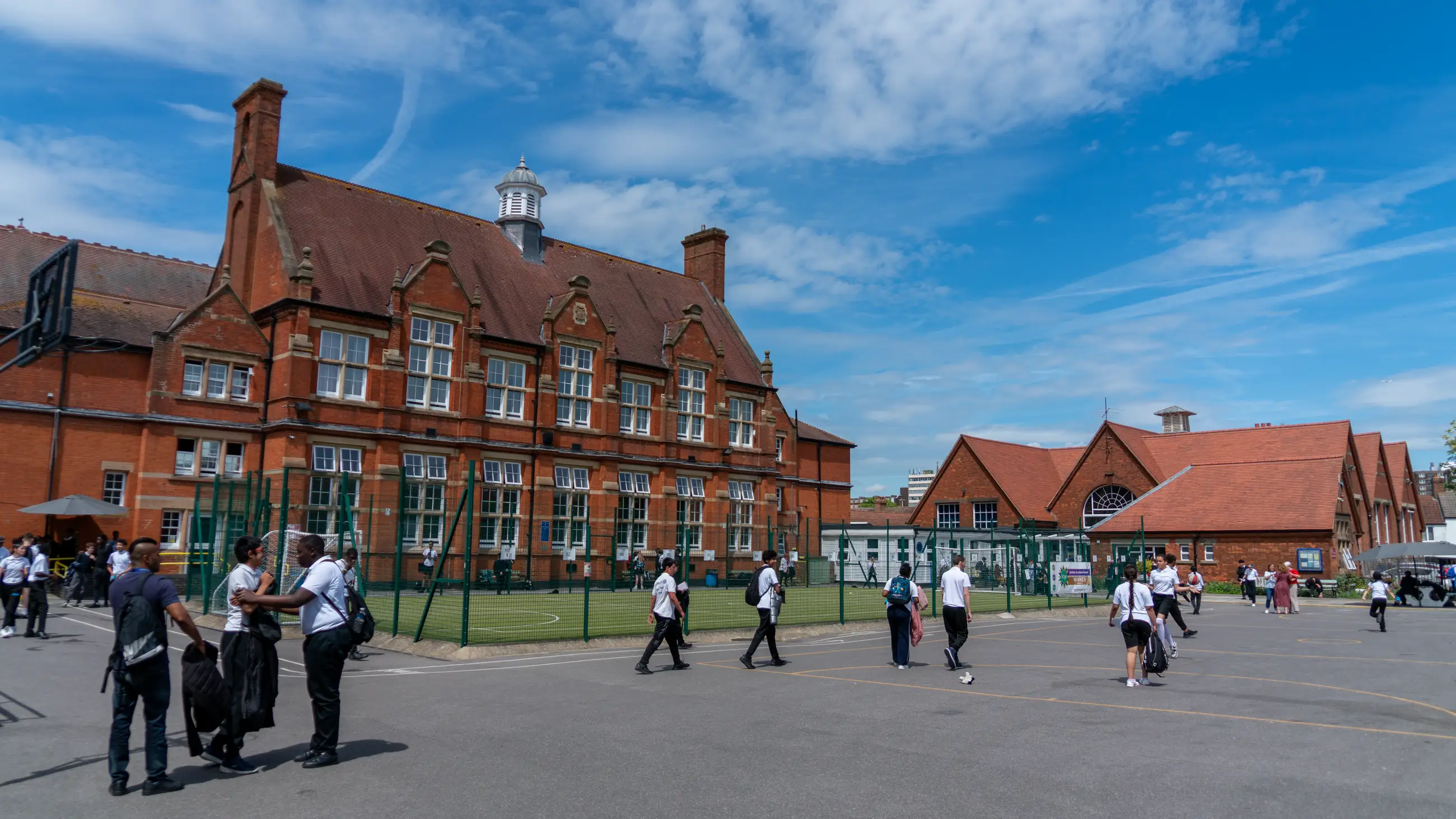 Students crossing the playground in front of the school's red-brick Victorian building on a sunny day.
