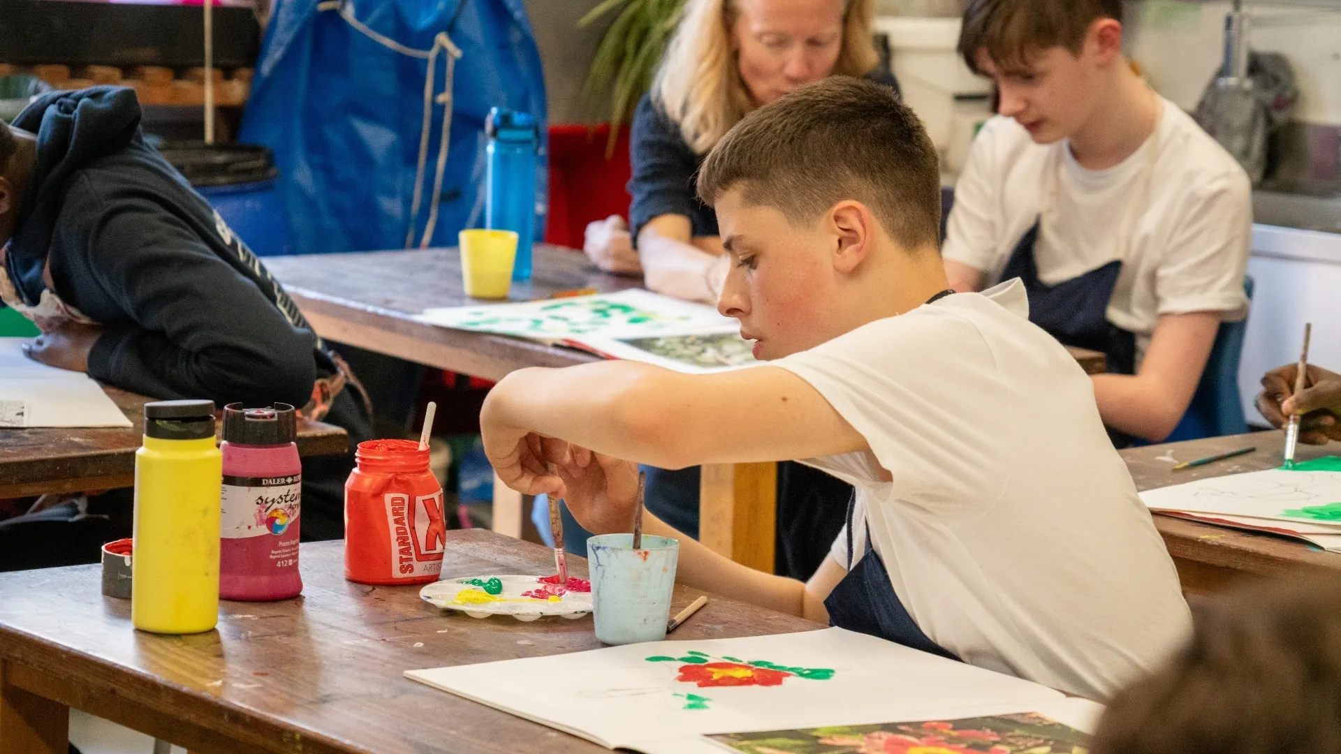 A student in an art lesson mixing red paint on a palette, classmates and a member of staff working at the tables around them.