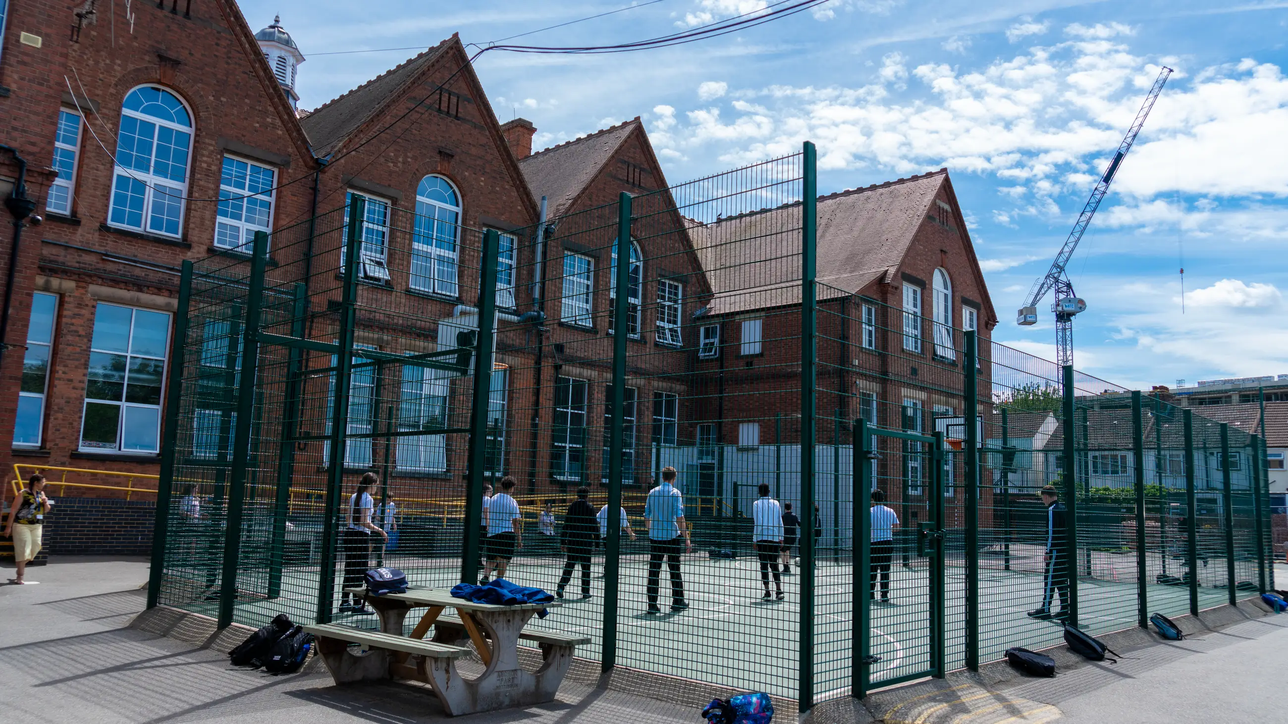 The school’s red-brick building behind the green-fenced ball court, with students playing at break.