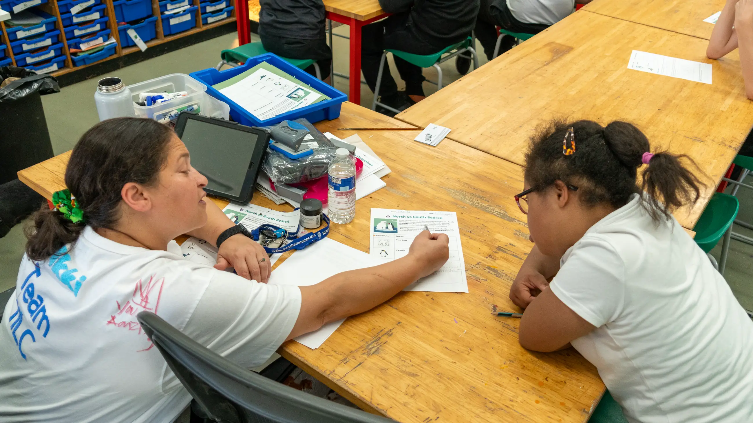 A member of staff working at a classroom table with two students, worksheets and a tablet in front of them.