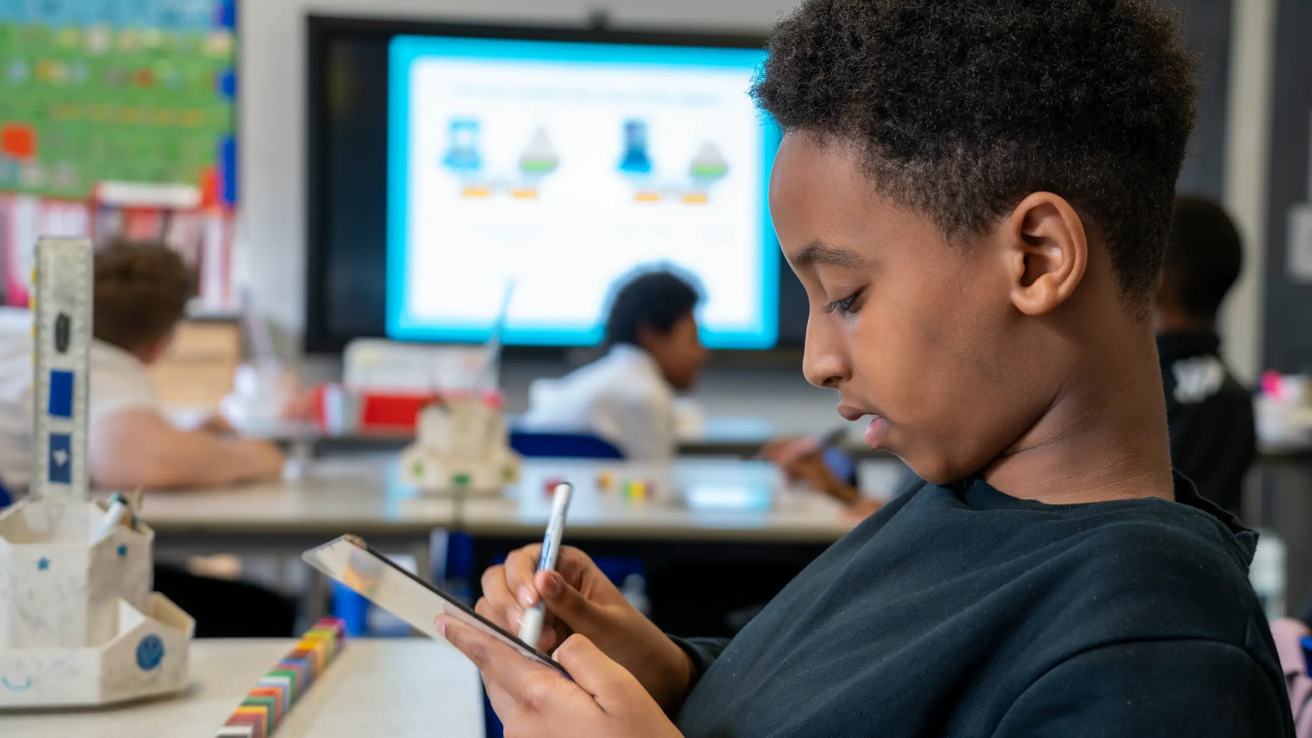 A student using a tablet in a classroom, an interactive screen behind.