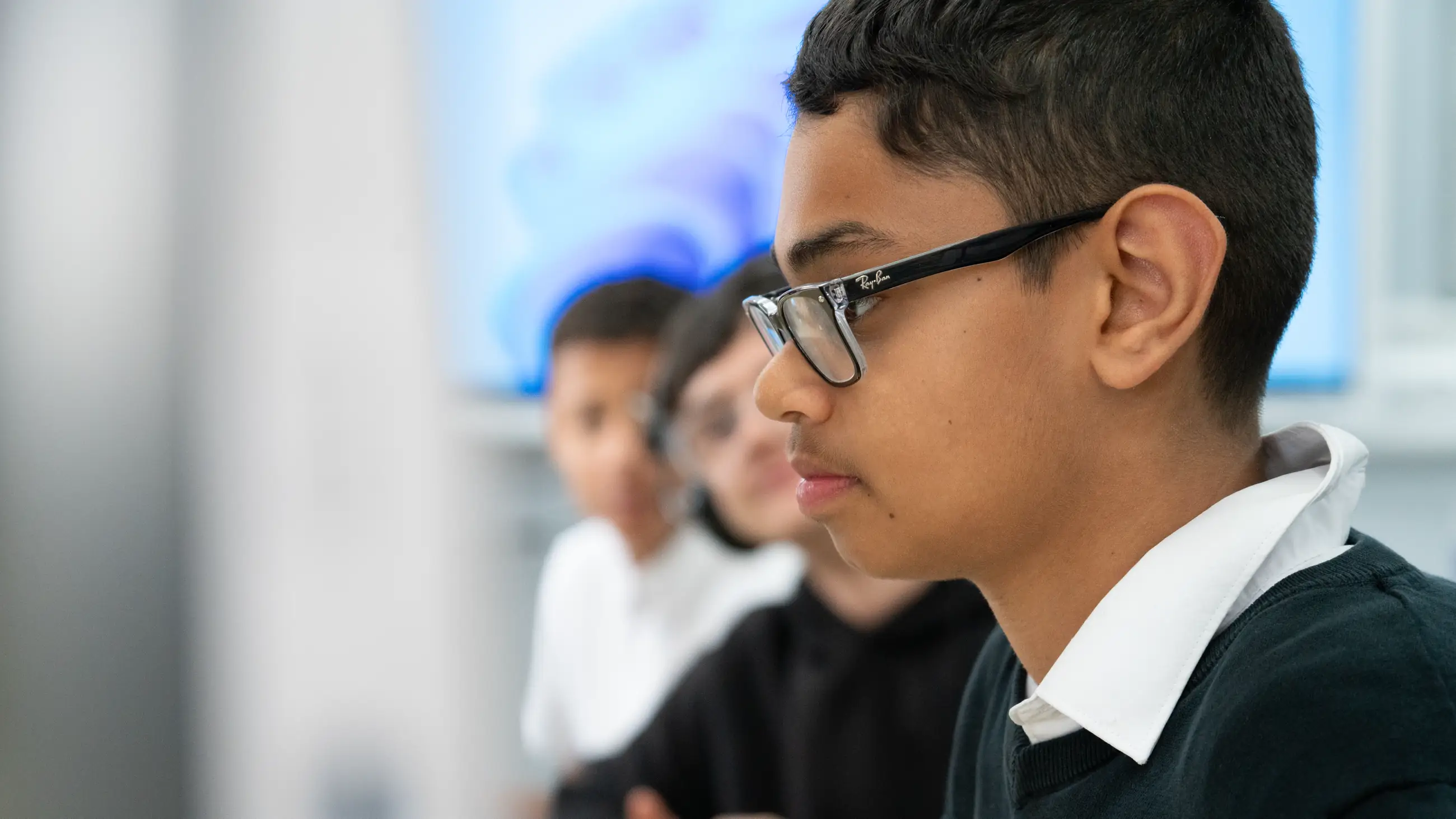 A student in school uniform and glasses concentrating during a lesson, classmates behind.