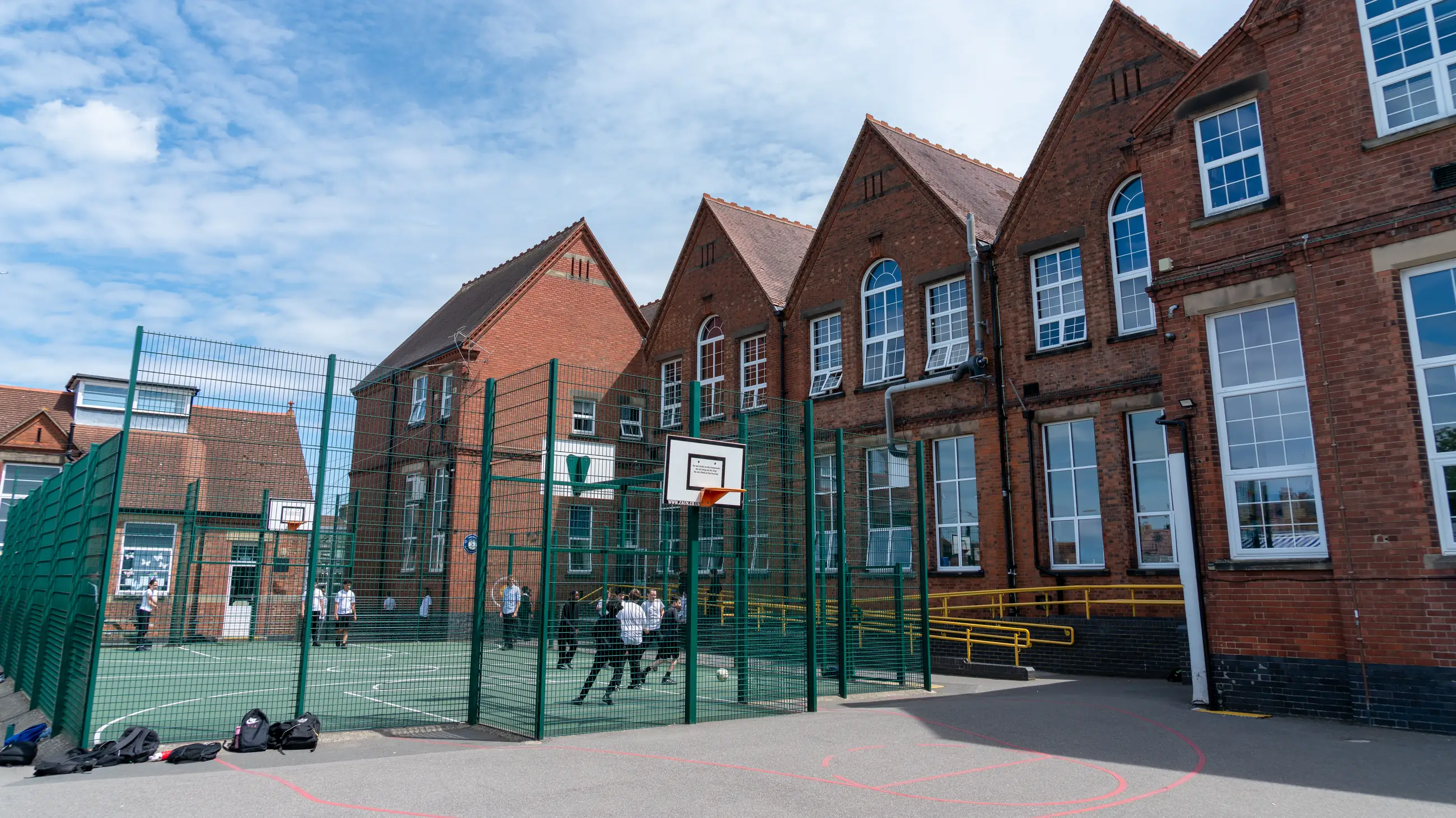 The school’s red-brick Victorian frontage seen across the fenced ball court, with students playing football.