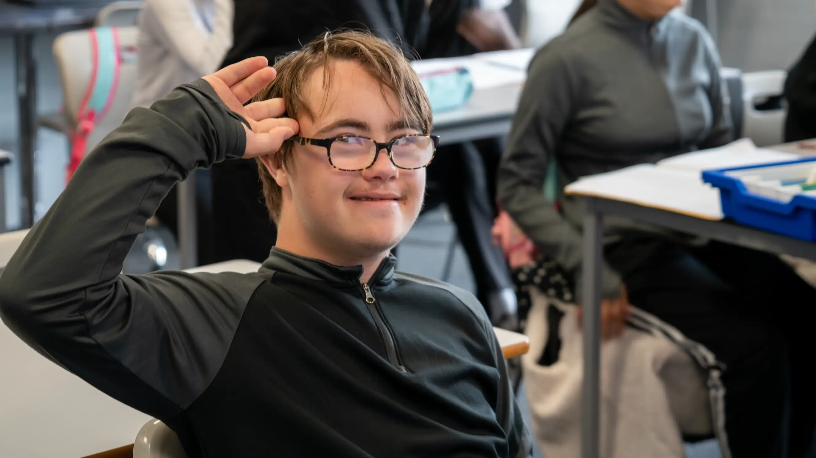 A student sitting back at a classroom desk, smiling towards the camera.