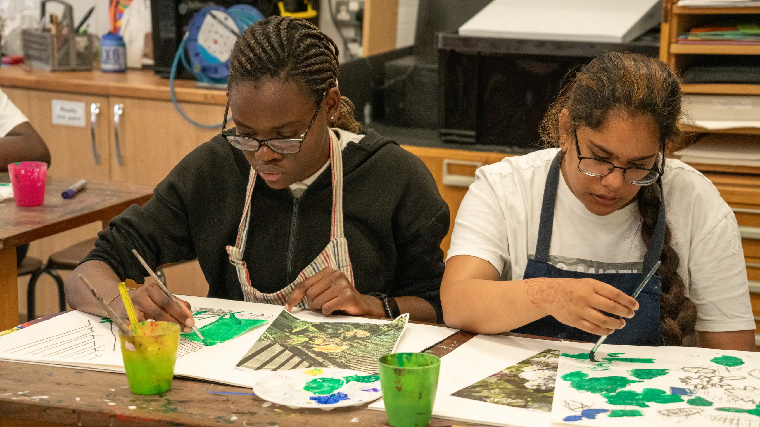Two students painting at a table, brushes and pots of paint in front of them.