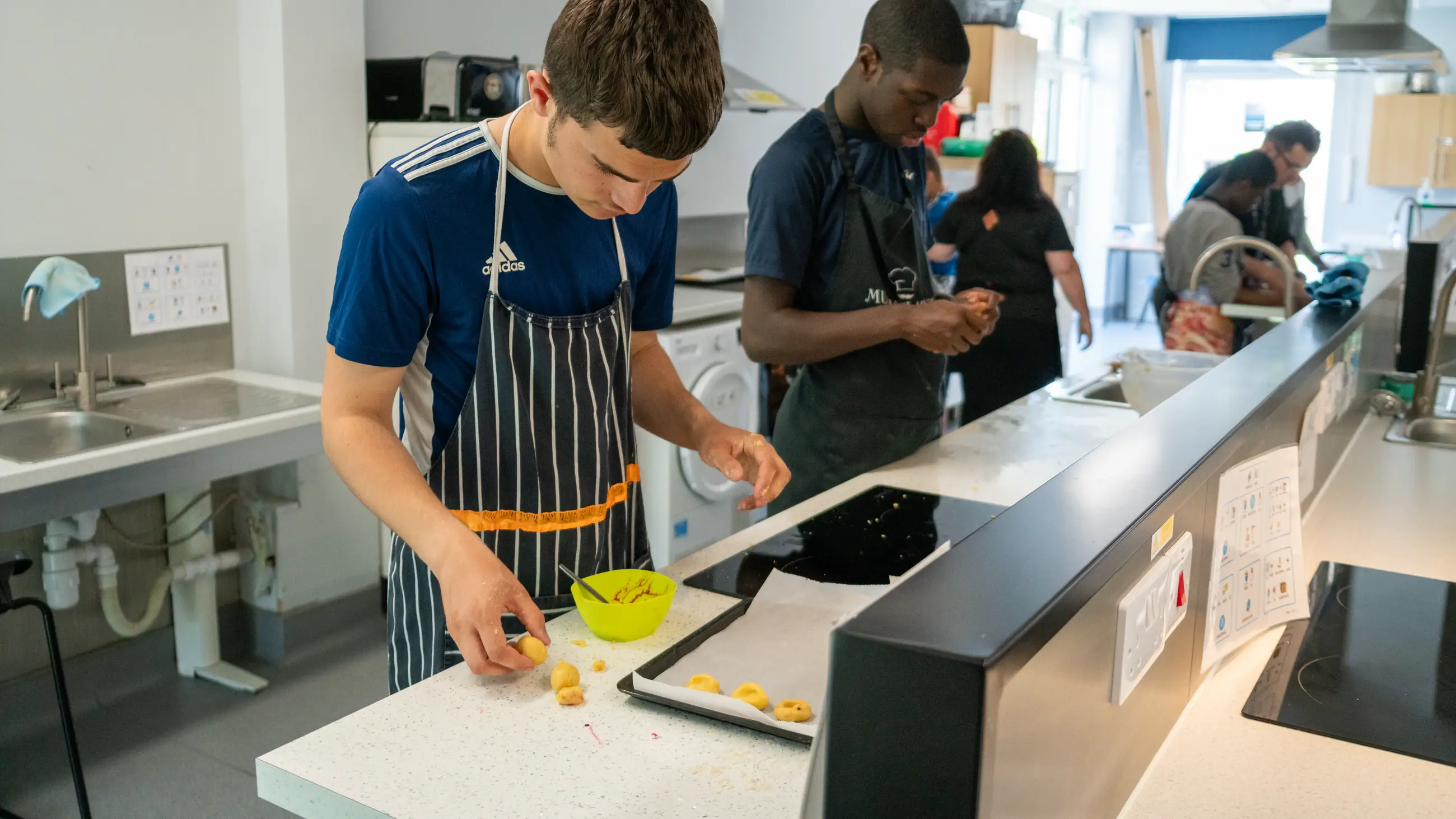Two students in aprons preparing food at a kitchen counter, a bowl and baking tray in front of them.