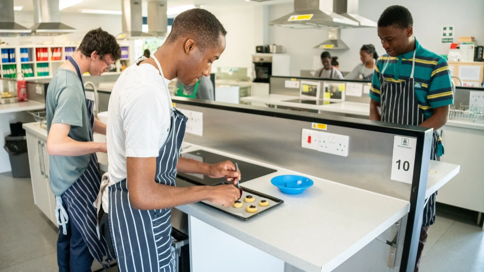Students in aprons at kitchen workstations, one arranging biscuits on a baking tray.