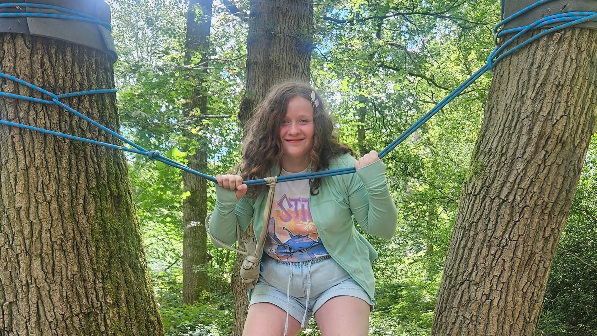 A student smiling on a rope bridge strung between two trees in the woods at Forest School.