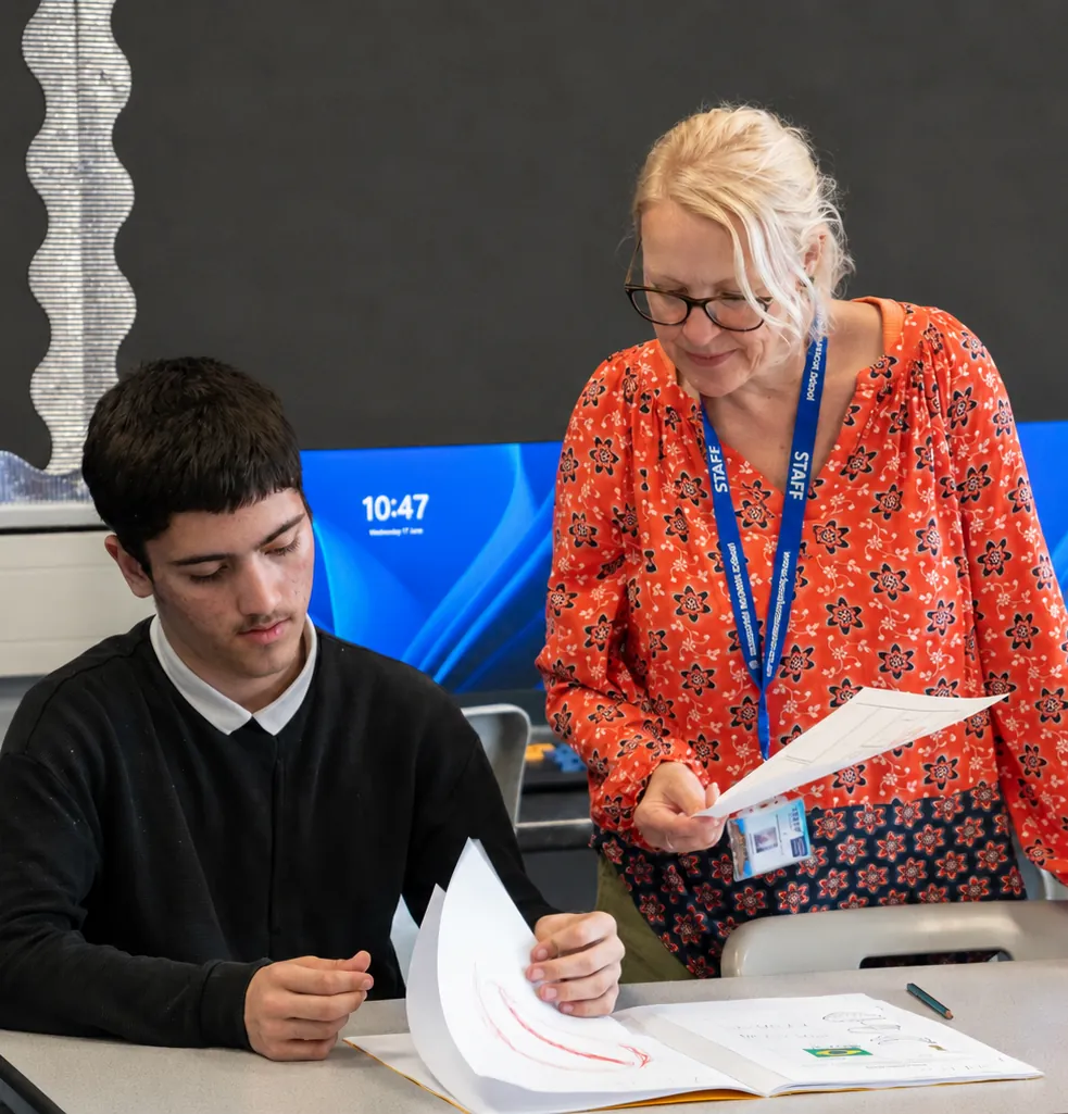 A member of staff standing beside a student at a desk in an ICT room, the two of them looking at his work together.