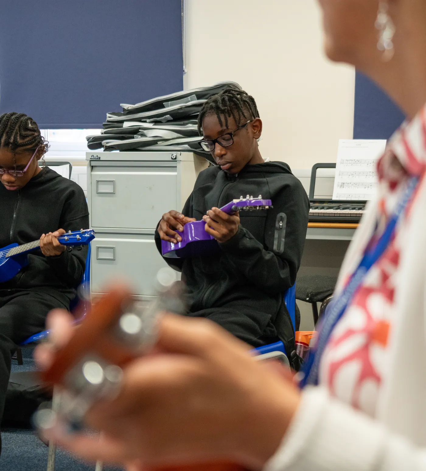 Two students in a music lesson playing ukuleles, a keyboard and sheet music on the table behind them.