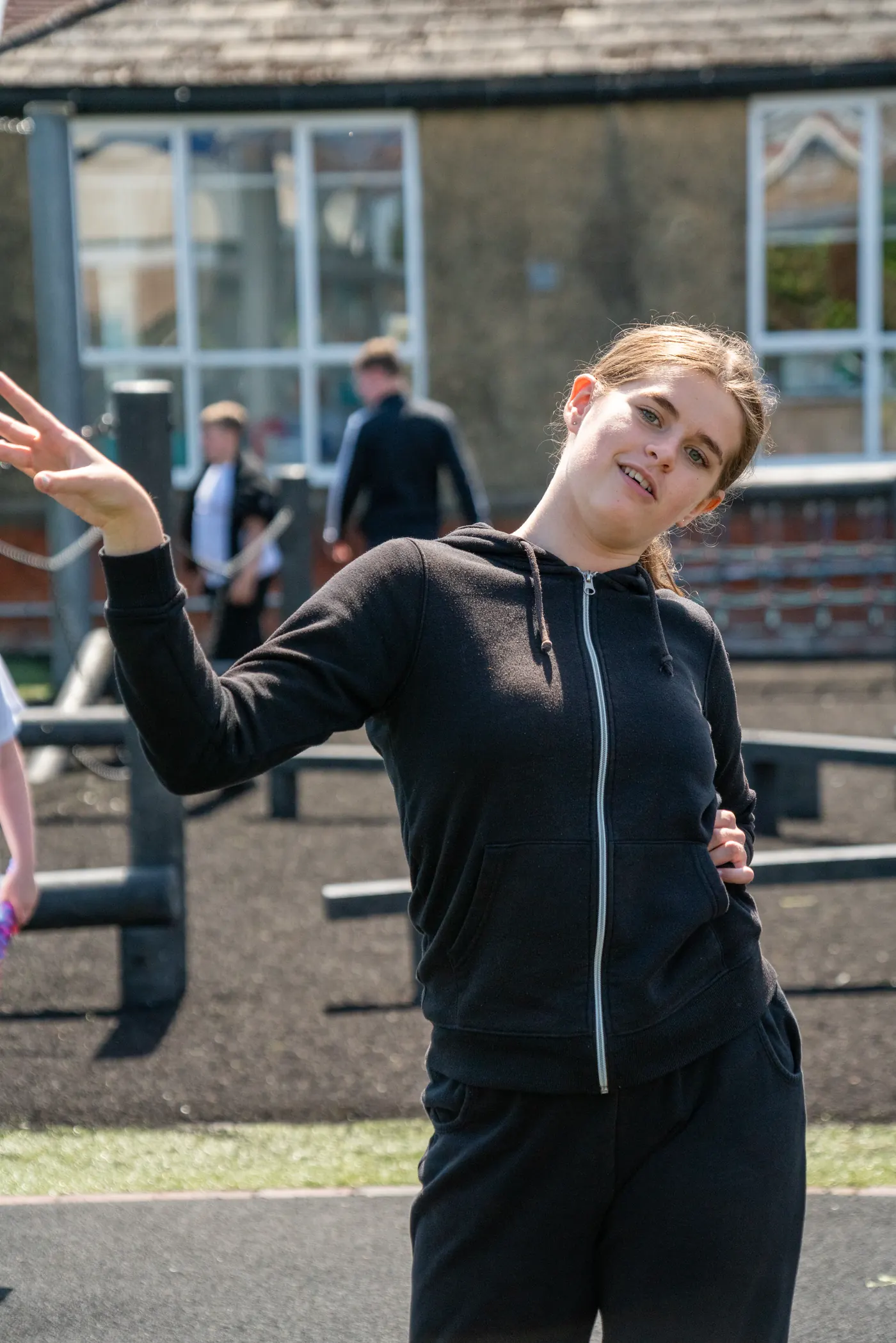 A student standing on the playground with one arm raised, classmates and the school building behind them.
