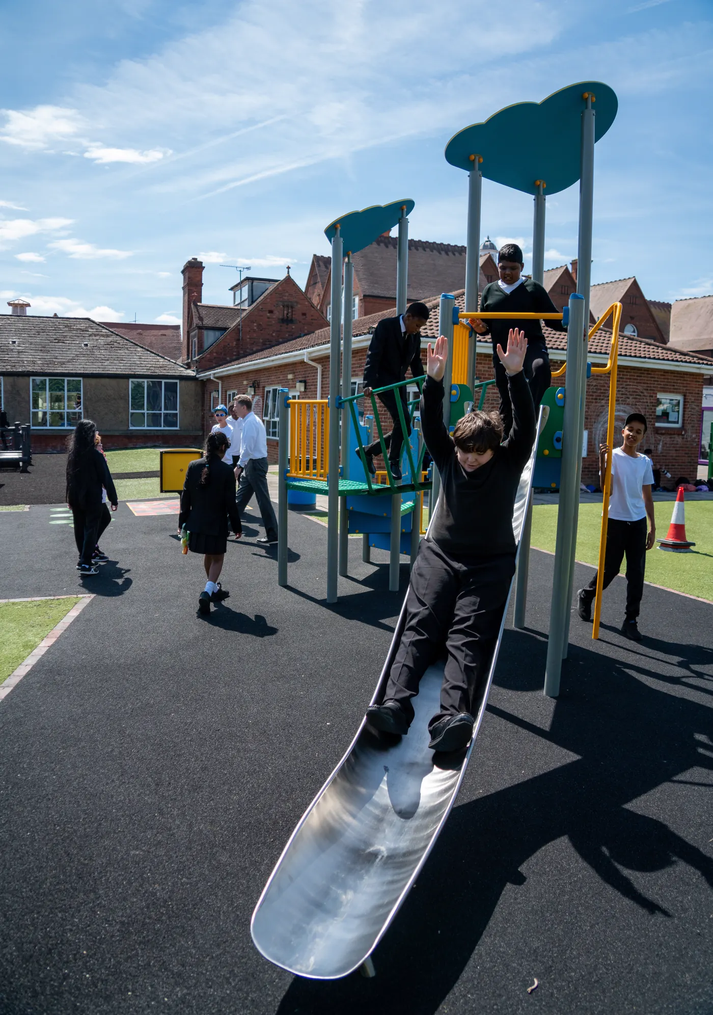 Students playing on the school playground, one coming down a slide with their arms in the air and others on the climbing frame behind.