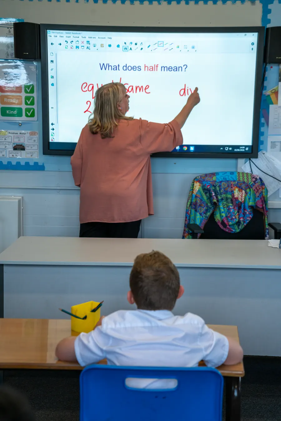 A teacher writing at an interactive whiteboard reading “What does half mean?” while a student watches from a desk.